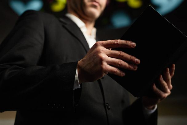 Close-up van een volwassen man in zwart pak die de bijbel leest en in de kerk staat - Foto, afbeelding