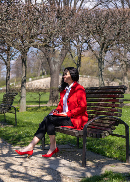 Κομψό επιχειρηματίας κάθεται σε ένα παγκάκι πάρκο και γράφει σε ένα σημειωματάριο. Η κυρία φοράει κόκκινο μανδύα, άσπρη μπλούζα και κόκκινα παπούτσια.. - Φωτογραφία, εικόνα