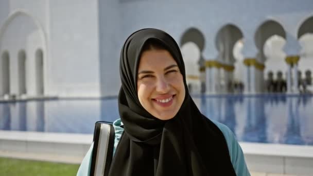 Μια χαμογελαστή γυναίκα με χιτζάμπ σε ένα τζαμί Abu dhabi με παραδοσιακή ισλαμική αρχιτεκτονική στο παρασκήνιο. - Πλάνα, βίντεο