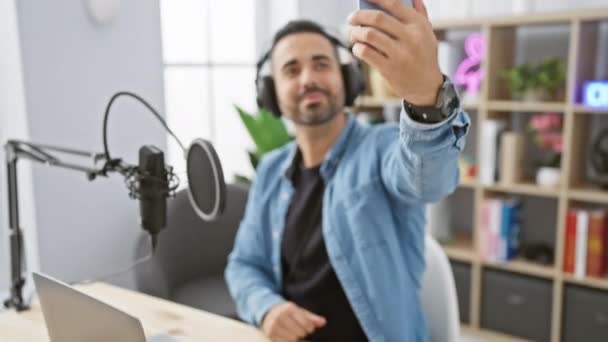 Hombre hispano guapo con barba sonriendo y tomando selfie en el estudio de radio usando smartphone y usando auriculares. - Metraje, vídeo