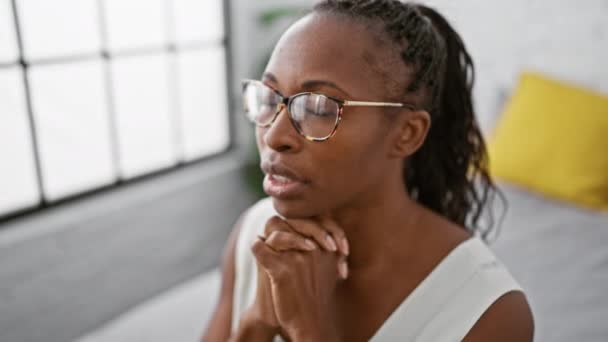 Contemplativa donna afroamericana con occhiali e capelli ricci in una luminosa sala interna - Filmati, video