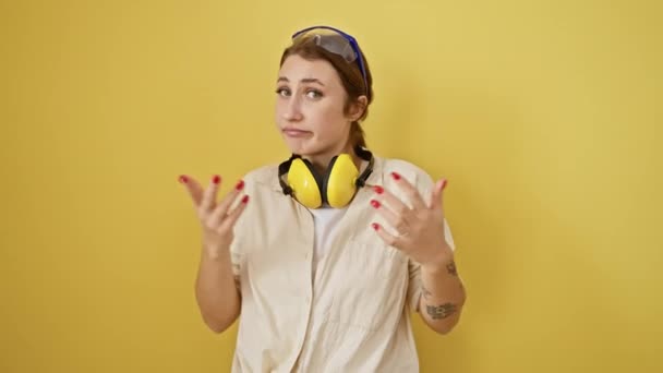 Baffled νεαρή μελαχρινή κοπέλα σε γυαλιά ασφαλείας και ακουστικά μείωσης του θορύβου με μια ανίδεη έκφραση, σηκώνοντας τα χέρια σε σύγχυση σε απομονωμένο κίτρινο φόντο - Πλάνα, βίντεο