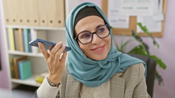 Mujer sonriente usando hijab usando teléfono inteligente en su oficina con fondo de estantería - Imágenes, Vídeo