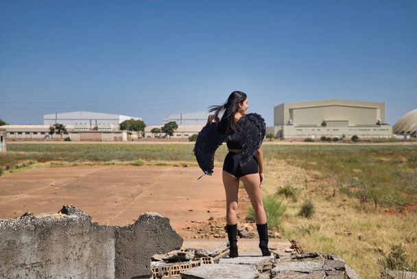 South American woman, young, beautiful, brunette with lingerie and black wings, posing on her back with the wind ruffling her hair amidst the ruins of a building. Concept angels, beauty, costumes. - Photo, Image