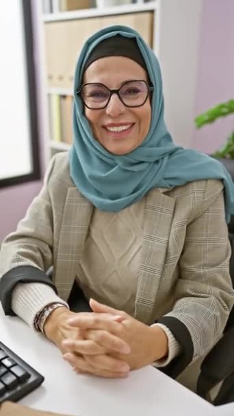Középkorú nő szemüveggel és hidzsáb mosollyal az íróasztalán, magabiztosságot és professzionalizmust árasztva. - Felvétel, videó