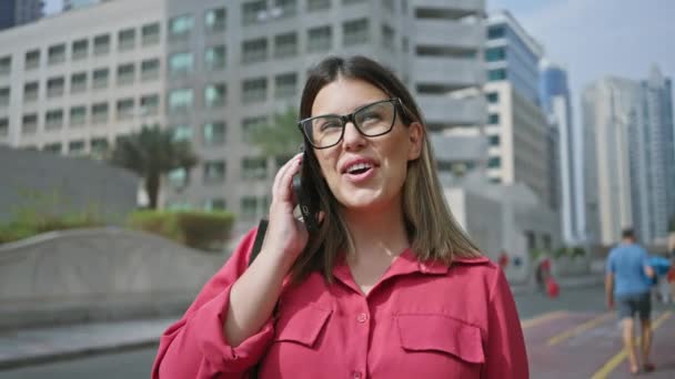 Glimlachende vrouw draagt bril praten op smartphone in dubai tegen moderne wolkenkrabbers achtergrond. - Video