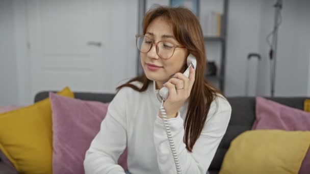 Een jonge vrouw met een bril bezig met een telefoongesprek terwijl ze comfortabel in haar moderne woonkamer zit. - Video