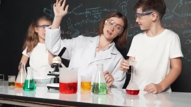 Běloch míchá chemickou tekutinu, zatímco učitel dává rady. Profesionální instruktor v laboratorním obleku, který hledá různorodého studenta u stolu s kádinkou plnou barevného roztoku. Erudice. - Záběry, video