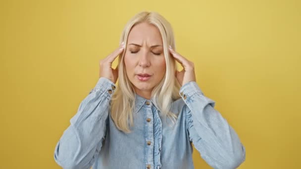 Stressed young woman in denim shirt feeling headache amidst a yellow isolated backdrop. blonde beauty suffering from migraine, standing with hand on head. - Footage, Video