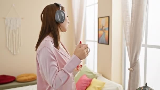 Mladá žena má ráda hudbu se sluchátky v útulné ložnici s šálkem a barevnými polštáři. - Záběry, video