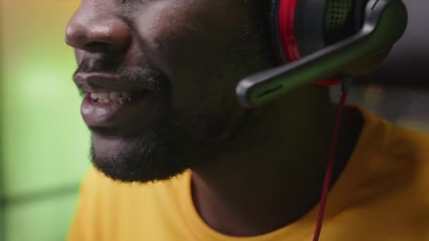 Κοντινό πλάνο του νεαρού μαύρου αρσενικού παίκτη που φοράει ακουστικά με μικρόφωνο που επικοινωνεί με τους παίκτες ενώ παίζει online βιντεοπαιχνίδι στο δωμάτιο νέον φως τη νύχτα - Πλάνα, βίντεο