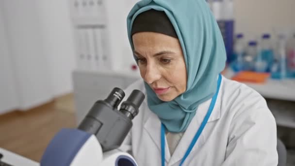 Soustředěná vědkyně používající mikroskop v laboratoři, oblečená v hidžábu a laboratorním plášti. - Záběry, video