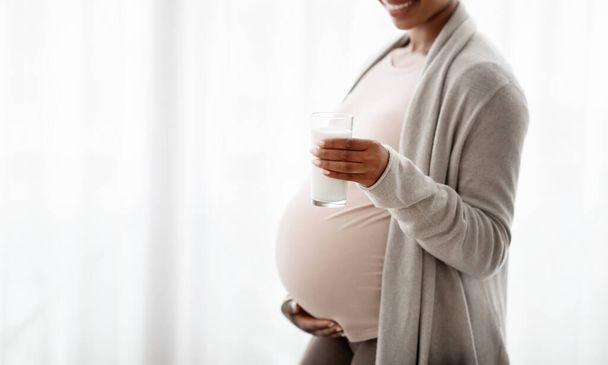 授乳と授乳のコンセプト。 自宅の窓の隣に立っている妊娠中の黒人女性のクロップ,彼女の腹を抱き,新鮮なミルクでグラスを保持し,コピースペースを持つパノラマ - 写真・画像