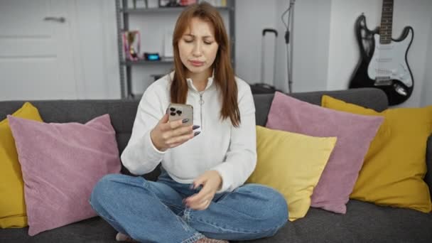 Een jonge vrouw checkt haar smartphone terwijl ze op een grijze bank zit met kleurrijke kussens in een gezellige woonkamer. - Video