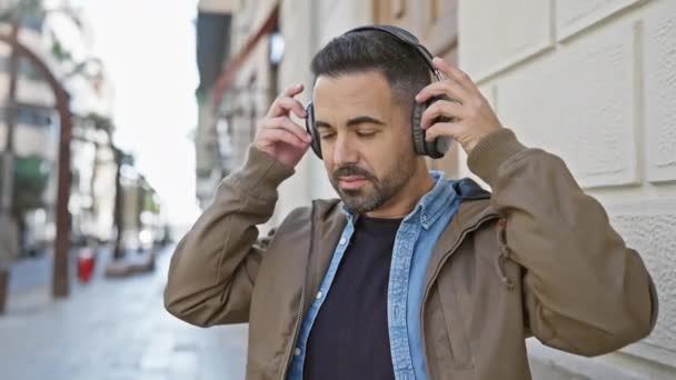 Een bebaarde jonge Latijns-Amerikaanse man met een koptelefoon staat in een straat in de stad en straalt stedelijke stijl en zelfvertrouwen uit.. - Video