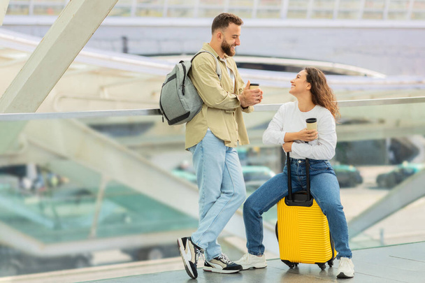 Feliz joven pareja divirtiéndose mientras espera vuelo en el aeropuerto, alegre europeo hombre y mujer bebiendo café para llevar y charlando, riendo y disfrutando de tiempo juntos, dama sentada en la maleta - Foto, imagen
