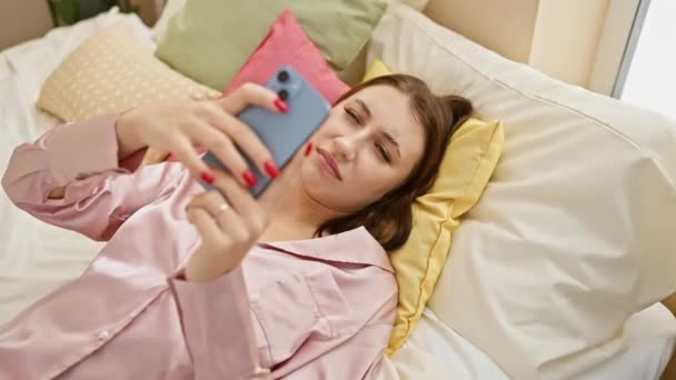 Μια νεαρή γυναίκα σε ένα ροζ πιτζάμα ξαπλωμένη άνετα στο κρεβάτι, ενώ χρησιμοποιώντας ένα smartphone σε ένα άνετο υπνοδωμάτιο ρύθμιση. - Πλάνα, βίντεο