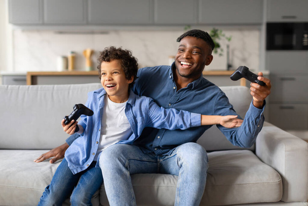 Un duo père-fils enthousiaste rit joyeusement en jouant ensemble à des jeux vidéo sur un canapé - Photo, image