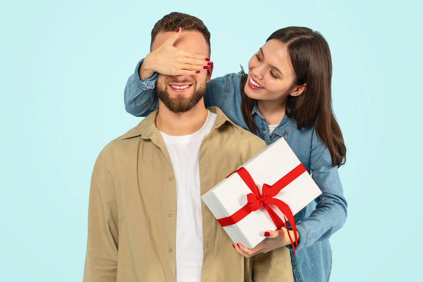 Young woman covers her boyfriends eyes surprising him with a birthday present against blue backdrop, posing with wrapped gift box, celebrating Valentines day or romantic relationship anniversary - Photo, Image