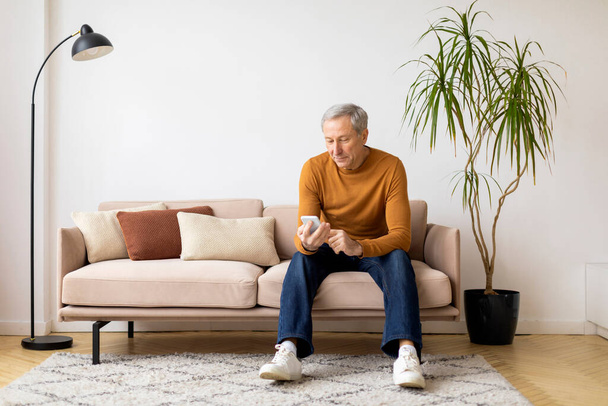 Vanhempi mies rento asu istuu sohvalla puhelimen kanssa, näyttää sitoutuneelta ja mukavalta kotiympäristössä. - Valokuva, kuva