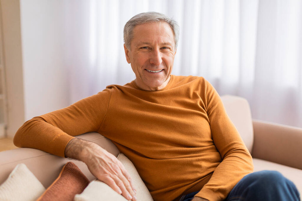 Confortable gentleman plus âgé se prélassant sur un canapé, souriant largement dans une pièce avec une sensation légère et aérée - Photo, image