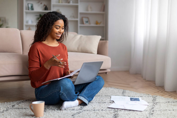 Hymyilevä nuori musta nainen, joka tekee videopuhelun kannettavalla tietokoneella kotona, istuu lattialla papereiden ympäröimänä valoisassa olohuoneessa, freelancer-nainen, joka tekee etäkonferenssin kotoa. - Valokuva, kuva