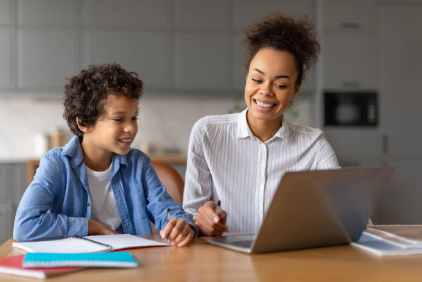 Een moeder en haar jonge zoon glimlachen terwijl ze aan een tafel zitten en samenwerken op een laptop, wat een thuisopleiding of huiswerksessie impliceert. - Foto, afbeelding