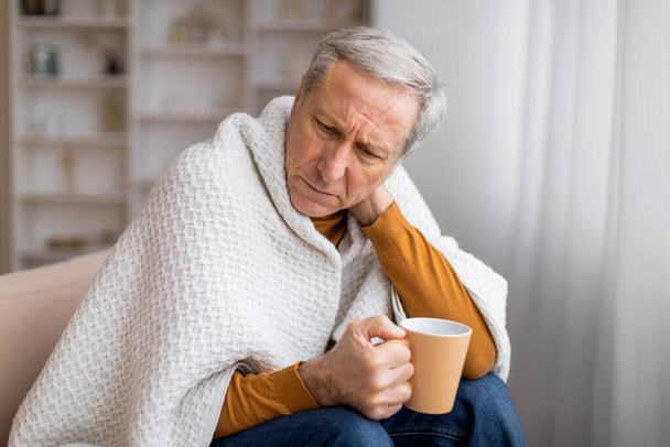 Enfermo mayor envuelto en una manta mientras sostiene una taza, sentado en el sofá, ilustrando problemas de salud o recuperación - Foto, imagen