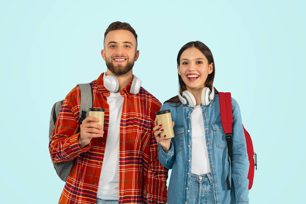 Fröhliches Studentenpaar posiert mit Rucksack und Kaffee zum Mitnehmen im Atelier, repräsentiert Freundschaft und Zweisamkeit in ihrer Studienzeit, steht lächelnd in die Kamera, blauer Hintergrund - Foto, Bild