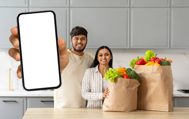 Onnellinen nuori intialainen puolisot tilata ostoksia verkossa, osoittaa paperipussi täynnä ruokaa ja älypuhelin valkoinen tyhjä näyttö mockup kopioi tilaa toimitus mobiilisovellus, kodikas keittiö sisustus - Valokuva, kuva