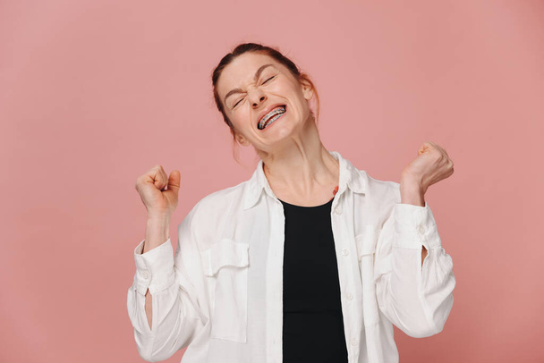 Moderne vrolijke vrouw glimlachen met beugels gebalde vuisten tonen winnen op roze achtergrond - Foto, afbeelding