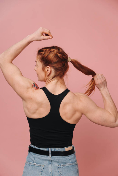 Спортивная мускулистая женщина в футболке и джинсах показывает сильную спину на розовом фоне - Фото, изображение