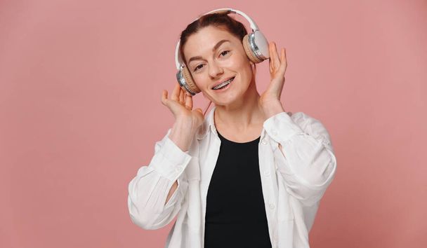 Modern woman smiling with braces on her teeth and listening to music in headphones on pink background - Photo, Image