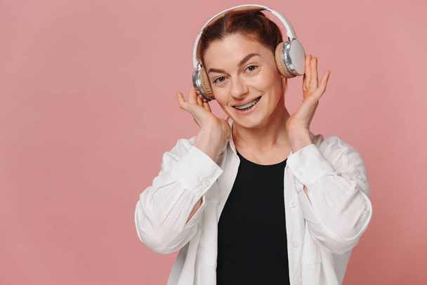 Сучасна жінка посміхається з брекетами на зубах і слухає музику в навушниках на рожевому фоні - Фото, зображення