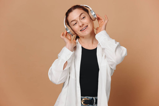 Mujer moderna sonriendo con frenillos en los dientes y escuchando música en auriculares sobre un fondo beige - Foto, imagen
