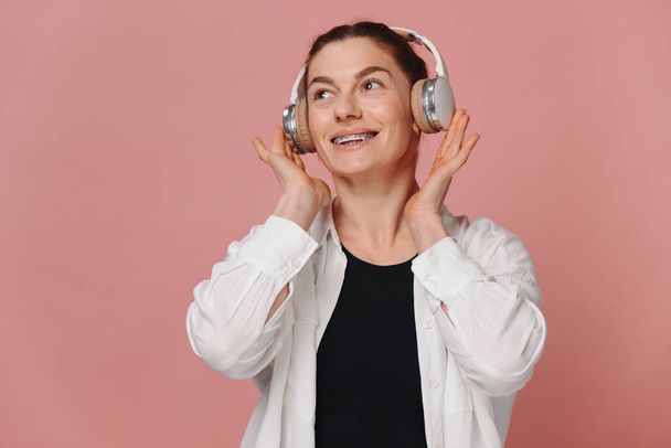Σύγχρονη γυναίκα χαμογελώντας με τιράντες στα δόντια της και ακούγοντας μουσική σε ακουστικά σε ροζ φόντο - Φωτογραφία, εικόνα