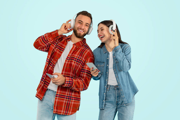 Smiling Caucasian Couple Disfrutando de la música en línea en los teléfonos, usando auriculares inalámbricos, escuchando canciones favoritas en los auriculares, de pie en el fondo del estudio azul. Concepto de aplicación musical - Foto, Imagen