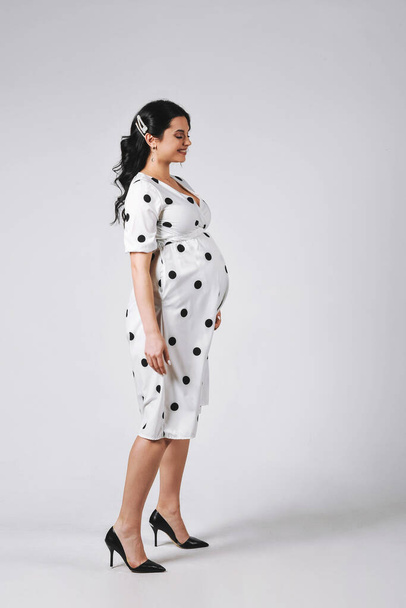 Retrato de estudio de mujer embarazada morena feliz en vestido de lunares blancos posando en un cyclorama. Copia espacio para texto. Bueno para papel pintado o revista de impresión - Foto, imagen