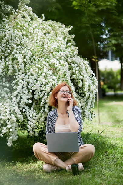 Happy young woman speaking by phone, using laptop in white flowering blooms. Remote work, chatting with friends, studying concept. - Photo, Image