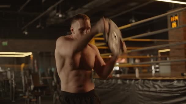 Un hombre desnudo está haciendo ejercicio en un gimnasio, levantando un plato mientras muestra su pecho muscular, abdomen y muslos, rodeado por la oscuridad de la habitación - Imágenes, Vídeo