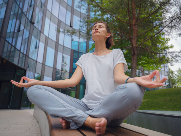 Sfatare, rilassare l'idea. pacifico dipendente freelance femminile che pratica esercizi di yoga mentre seduto in gruppo sotto grattacieli di vetro dopo una dura giornata di lavoro in ufficio, tenendosi per mano nel mudra - Foto, immagini