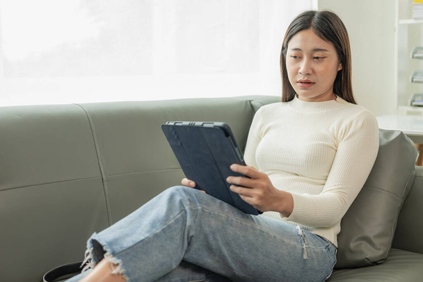 Ελκυστική νεαρή Ασιάτισσα που κάθεται σε γκρι καναπέ και χρησιμοποιεί ψηφιακή ταμπλέτα στο σαλόνι, στέλνοντας μήνυμα στο tablet. Χαμογελώντας νεαρή γυναίκα χρησιμοποιεί ένα tablet online μήνυμα chat Ψώνια online από το σπίτι - Φωτογραφία, εικόνα