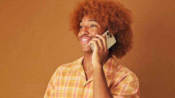 Smiling stylish curly guy speaks emotionally on mobile phone, isolated on brown background in the studio - Foto, immagini