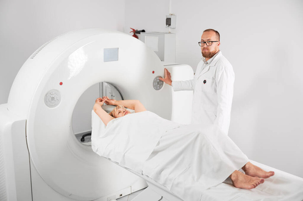 Tomografia medyczna lub rezonans magnetyczny. Mężczyzna radiolog naciska przycisk MRI, aby zbadać pacjentkę, patrząc w kamerę. - Zdjęcie, obraz