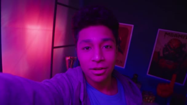Handheld POV of Biracial teenage gamer boy speaking on camera while recording video blog in neon light room at night - Footage, Video
