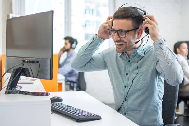 Hombre sonriente operador de servicio al cliente con sus auriculares profesionales y preparándose para trabajar frente a la computadora en su lugar de trabajo en el centro de llamadas - Foto, Imagen