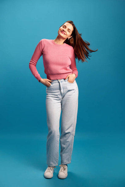 Relaxed and positive young woman in pink sweater and blue jeans cheerfully dancing and having fun against blue studio background. Concept of youth, lifestyle, casual fashion, human emotions - Foto, Imagem