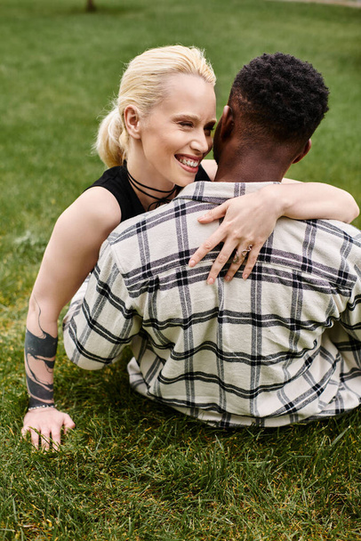 A happy multicultural couple, an African American man and a Caucasian woman, sharing a tender hug on the grass in a park. - Фото, изображение