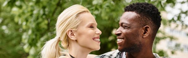 Un hombre afroamericano feliz está al lado de una mujer rubia al aire libre en un parque, sonriendo y conectando. - Foto, imagen