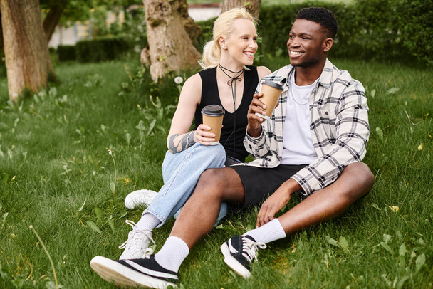 Wielokulturowa para, Afroamerykanin i biała kobieta, siedzą szczęśliwie razem na trawie w parku. - Zdjęcie, obraz